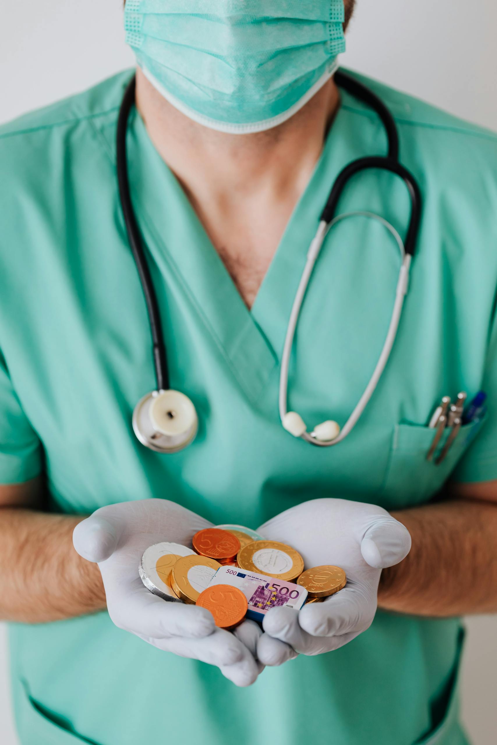 Anonymous male doctor wearing medical uniform mask and latex gloves holding coins and plastic euro while standing near white wall in clinic